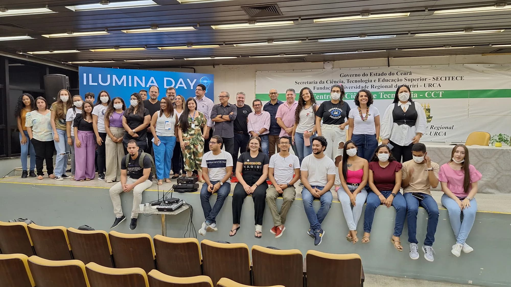 Pessoas da comunidade reunidas no palco de um auditório, com o painel Ilumina Day ao fundo