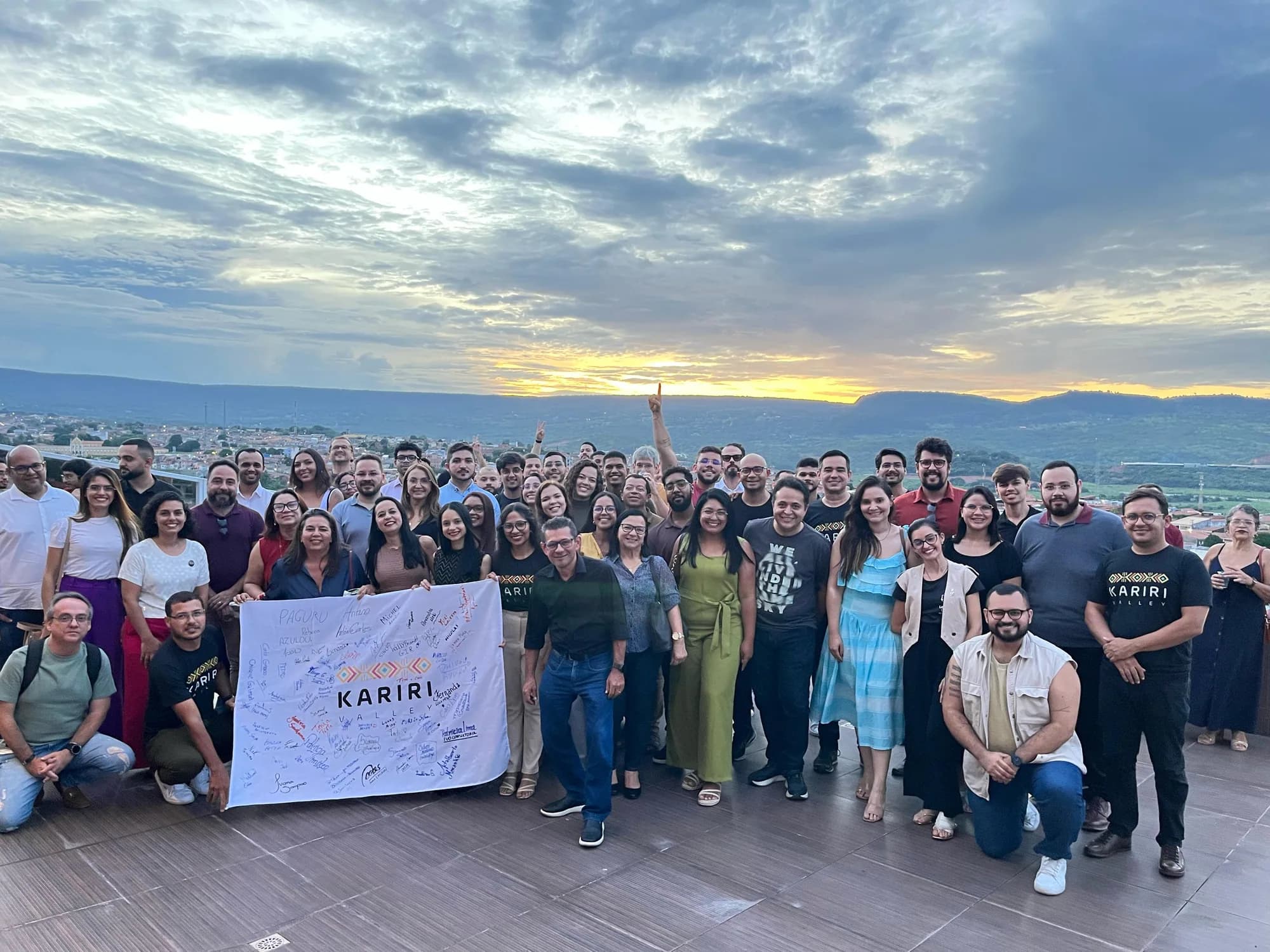 Grande grupo da comunidade segura a bandeira do Kariri Valley em uma varanda ao pôr do sol