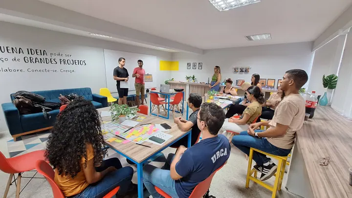 Participantes compartilham ideias em uma sala com mesas e materiais de trabalho