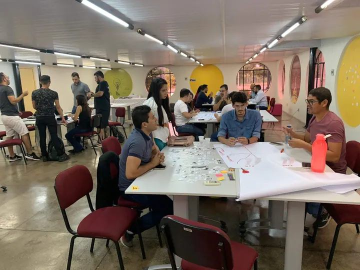 Participantes trabalham em grupos ao redor de mesas em uma sala de inovação