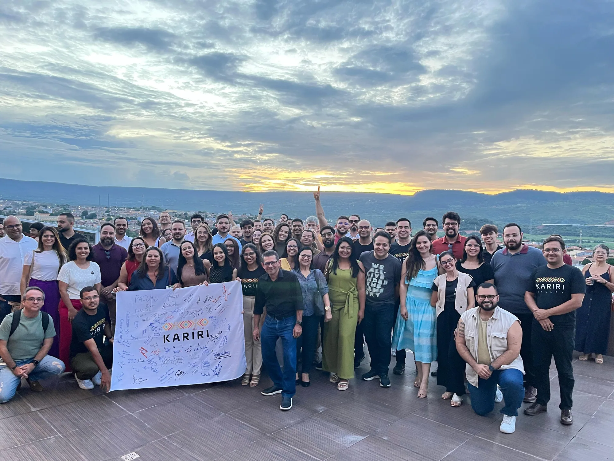 Grande grupo da comunidade segura a bandeira do Kariri Valley em uma varanda ao pôr do sol